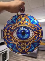 Evil eye acrylic suncatcher with blue, turquoise, orange and gold stained-glass-style design hanging by a window